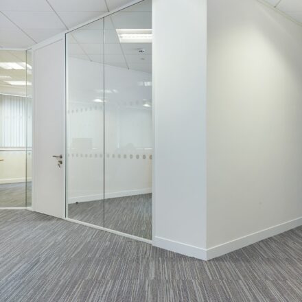 White corner partitioning with floor to ceiling glazed panels and grey carpet.