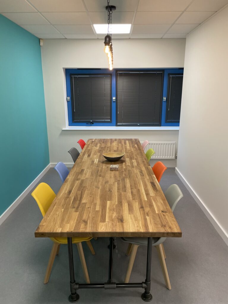 Large wooden table with bright coloured chairs, a turquoise feature wall and lighting over centre of table.