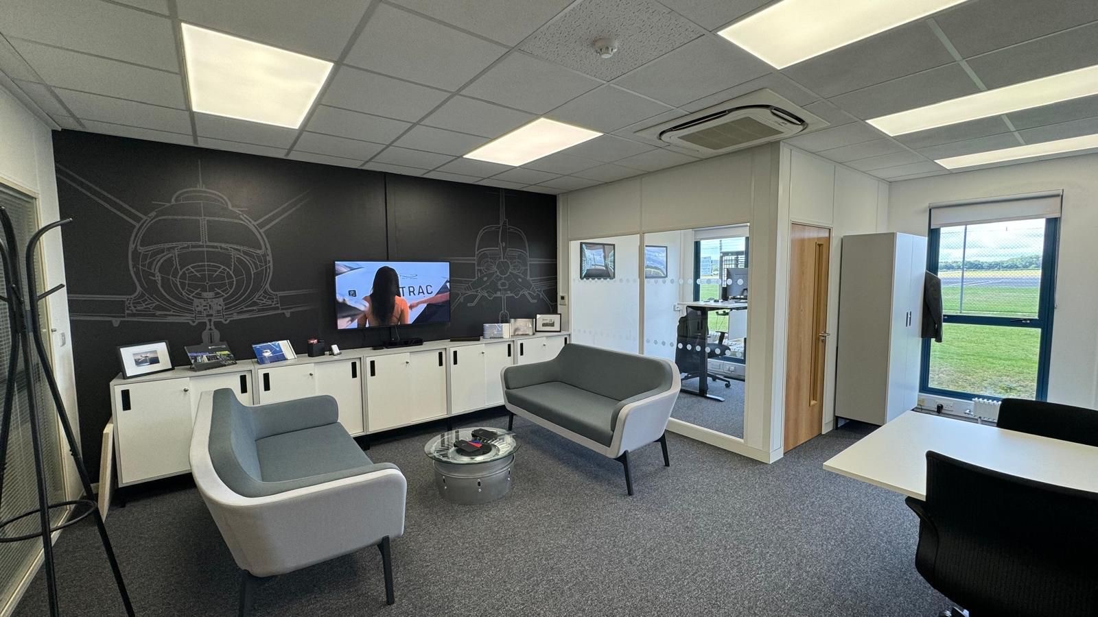 Black wall with aircraft graphics. Low level storage units and grey sofas in foreground. Corner office, with glazed wall.