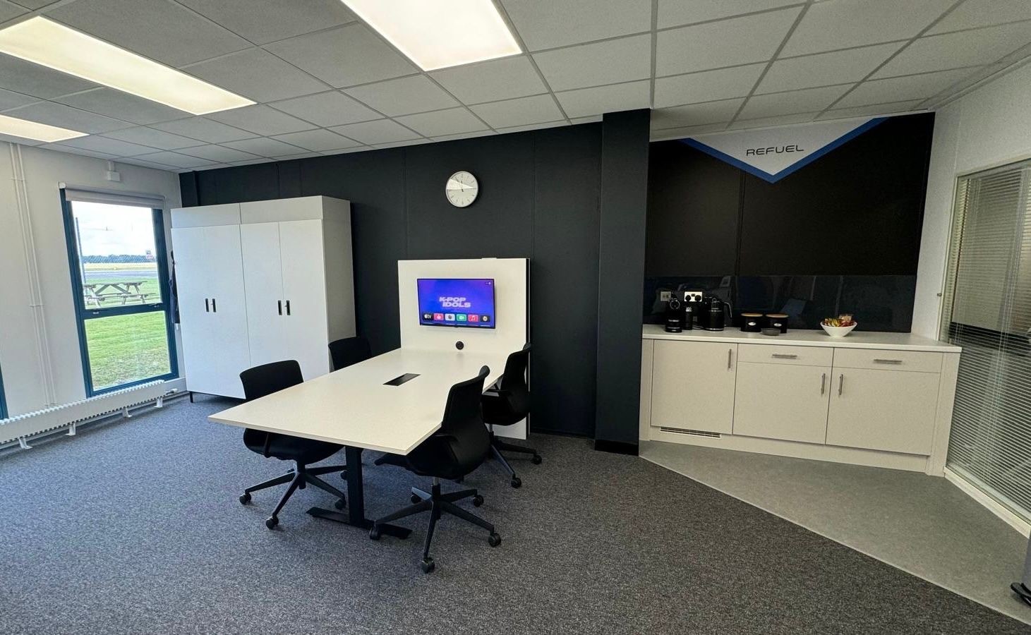 Black wall with large white storage unit and white meeting table in front. A small tea making area to the right of the image.