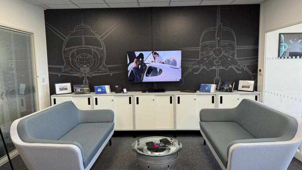 Black wall with aircraft graphics on, with TV mounted on wall in front. Grey sofas in front on low level white storage units and a small round coffee table.
